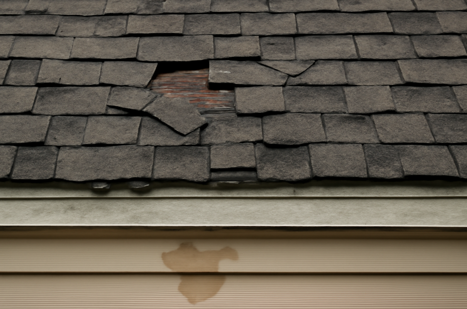 "Close-up view of a damaged roof with cracked and missing shingles, revealing exposed wood beneath. A water stain is visible on the beige siding below, indicating a roof leak. Moss and dirt accumulation on the shingles highlight the roof's aged condition."







